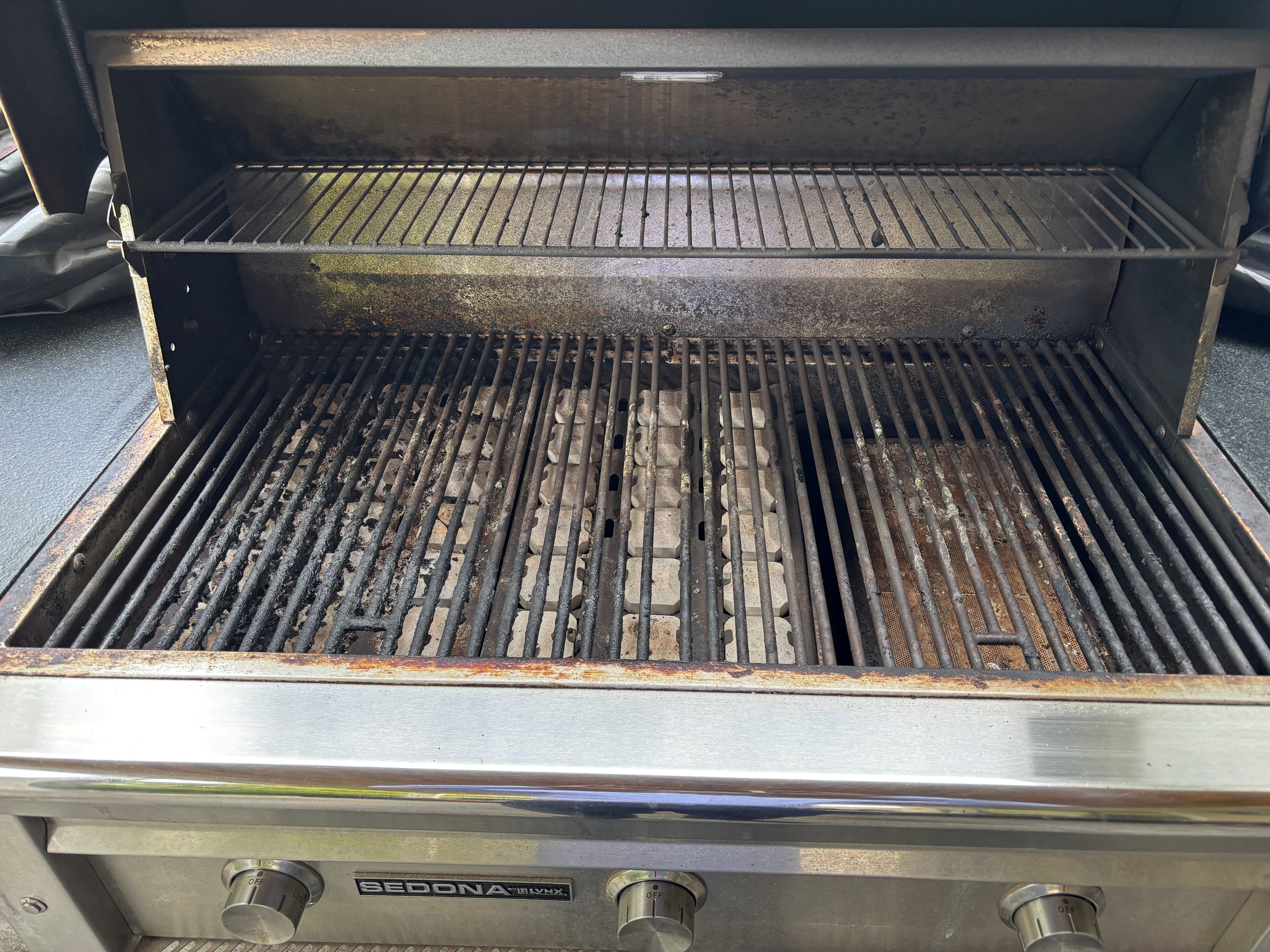 Sedona by Lynx gas grill before an Aspect deep clean, with soot-caked cooking grates, ash across the burner box and a grease-filmed warming rack